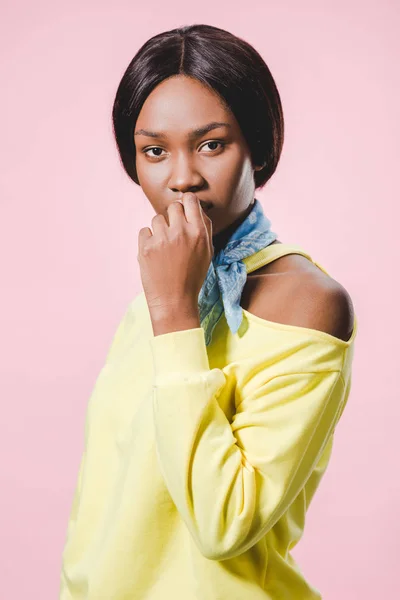 Mujer afroamericana pensativa en jersey amarillo y bufanda mirando a la cámara aislada en rosa - foto de stock