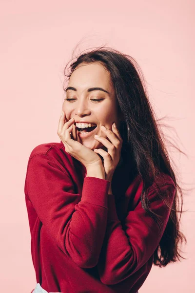 Привлекательная азиатка с закрытыми глазами в красном свитере, улыбающаяся изолированно на розовом — стоковое фото