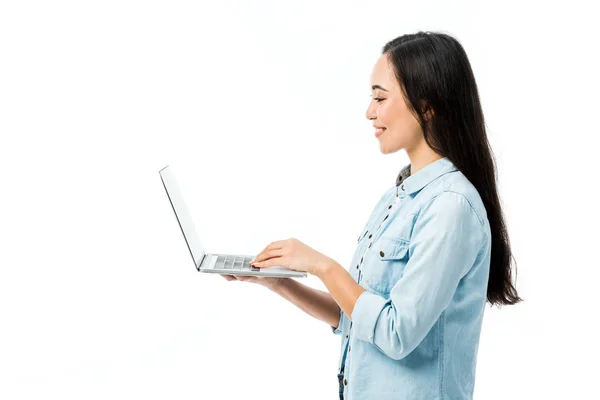 Vista laterale di attraente donna asiatica in camicia di jeans sorridente e tenendo il computer portatile isolato su bianco — Foto stock