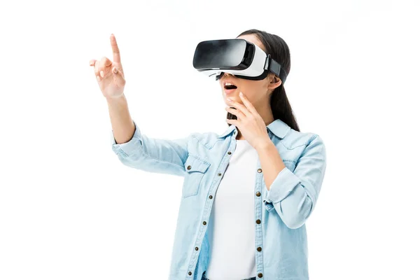 Donna scioccata in camicia di jeans con auricolare realtà virtuale punta con dito isolato su bianco — Foto stock