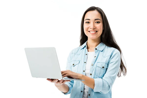 Atractiva mujer asiática en camisa de mezclilla sonriendo y sosteniendo portátil aislado en blanco - foto de stock