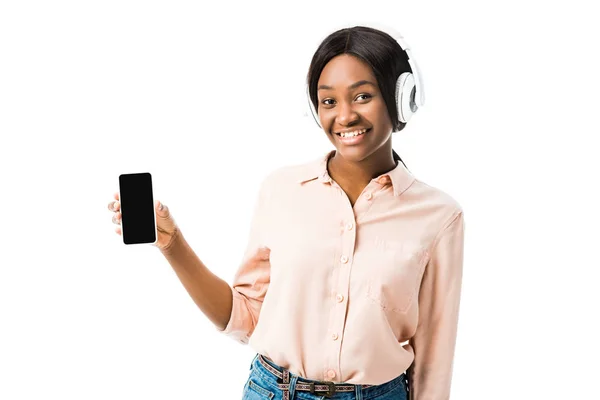 Усміхнена афроамериканська жінка в сорочці з навушниками тримає смартфон ізольовано на білому — стокове фото