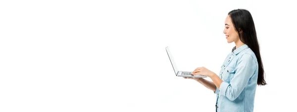 Tiro panorâmico de mulher asiática atraente em camisa jeans sorrindo e segurando laptop isolado no branco — Fotografia de Stock