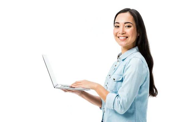 Atractiva mujer asiática en camisa de mezclilla sonriendo y sosteniendo portátil aislado en blanco - foto de stock