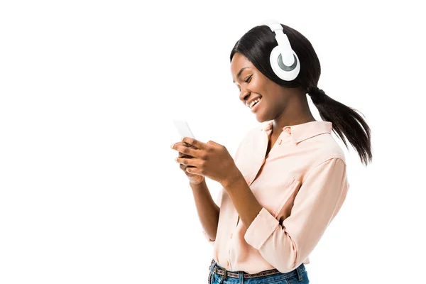 Улыбающаяся африканская американка в рубашке с наушниками, используя смартфон, изолированный на белом — стоковое фото