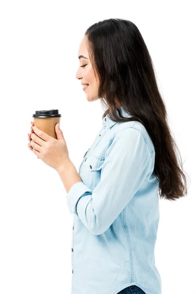 Вид сбоку привлекательной и улыбчивой женщины в джинсовой рубашке, держащей бумажный стаканчик, изолированный на белом — стоковое фото