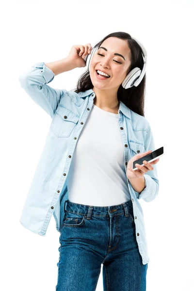 Atraente e sorridente asiático mulher no denim camisa ouvindo música e segurando smartphone isolado no branco — Fotografia de Stock