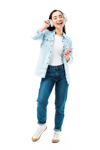 Attraktive und lächelnde asiatische Frau in Jeanshemd, die Musik hört und ihr Smartphone isoliert auf weiß hält — Stockfoto