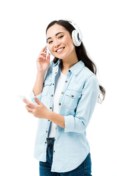 Atractiva y sonriente mujer asiática en camisa vaquera escuchando música y sosteniendo teléfono inteligente aislado en blanco — Stock Photo