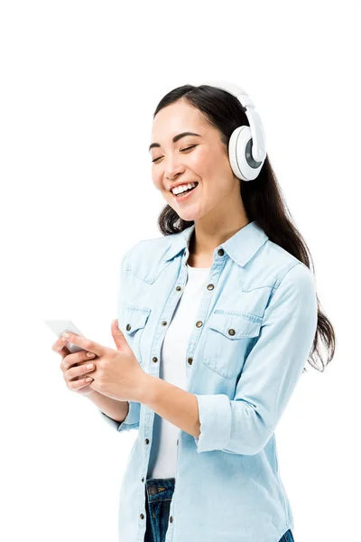 Attraktive und lächelnde asiatische Frau in Jeanshemd, die Musik hört und ihr Smartphone isoliert auf weiß hält — Stockfoto