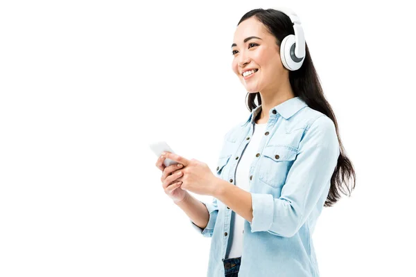 Atractiva y sonriente mujer asiática en camisa vaquera escuchando música y sosteniendo teléfono inteligente aislado en blanco - foto de stock