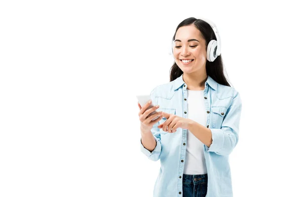 Attraente e sorridente asiatico donna in denim camicia ascolto musica e tenendo smartphone isolato su bianco — Foto stock