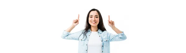 Colpo panoramico di attraente e sorridente donna asiatica in camicia di jeans che punta con le dita isolate sul bianco — Foto stock