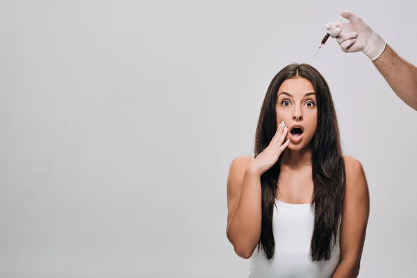 Cosmetologo in lattice guanti facendo mesoterapia del cuoio capelluto per scioccato bella donna con i capelli lunghi isolati su grigio — Foto stock