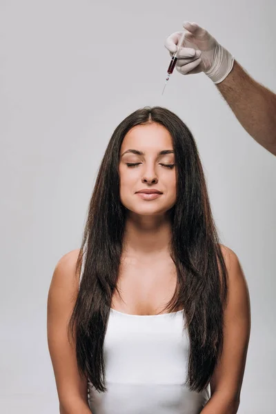 Cosmetologista em luvas de látex fazendo mesoterapia couro cabeludo para morena bela mulher com cabelos longos isolados em cinza — Fotografia de Stock