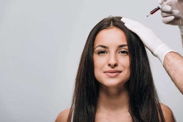 Cosmetologo in guanti di lattice facendo mesoterapia del cuoio capelluto per sorridere bella donna con i capelli lunghi isolati su grigio — Foto stock