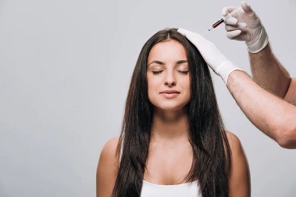 Kosmetikerin in Latex-Handschuhen macht Kopfhaut Mesotherapie für brünette schöne Frau mit langen Haaren und geschlossenen Augen isoliert auf grau — Stockfoto