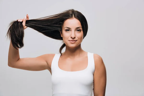 Sonriente morena hermosa mujer sosteniendo largo recta sano y brillante pelo aislado en gris - foto de stock