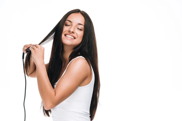 Morena sonriente hermosa mujer con los ojos cerrados alisando el pelo largo brillante con hierro plano aislado en blanco - foto de stock