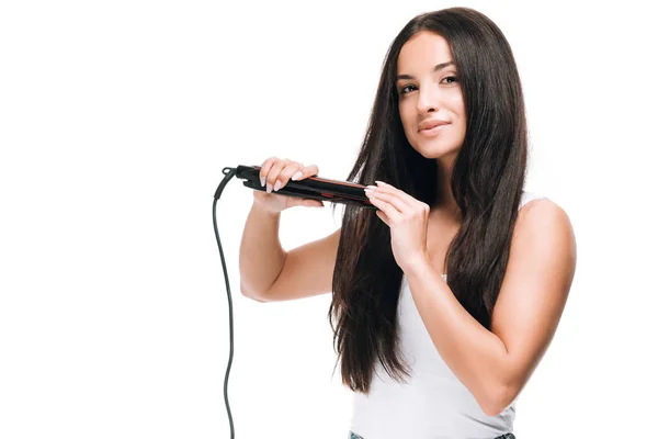 Bruna sorridente bella donna raddrizzamento lunghi capelli lucidi con ferro piatto isolato su bianco — Foto stock