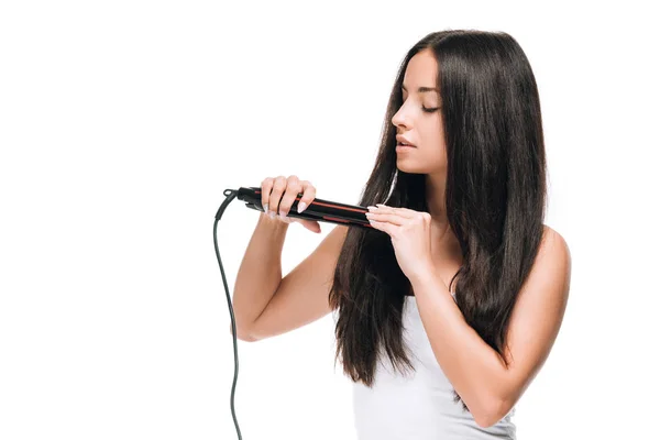 Brunette beautiful woman with closed eyes straightening long shiny hair with flat iron isolated on white — Stock Photo