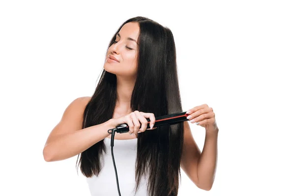 Smiling brunette beautiful woman straightening long shiny hair with flat iron isolated on white — Stock Photo