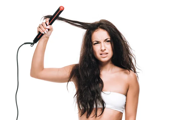 Confused brunette beautiful woman styling curly hair with flat iron isolated on white — Stock Photo