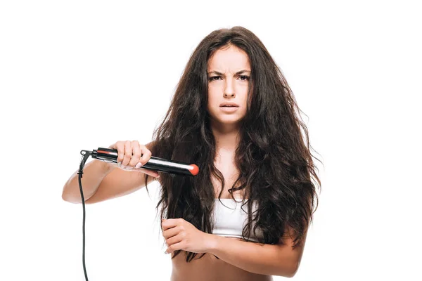 Stressed brunette beautiful woman styling curly hair with flat iron isolated on white — Stock Photo