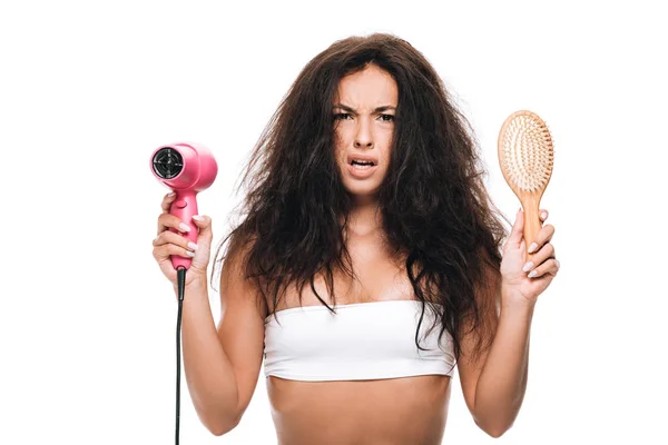 Inquiet brunette belle femme avec les cheveux bouclés tenant sèche-cheveux rose et peigne isolé sur blanc — Photo de stock