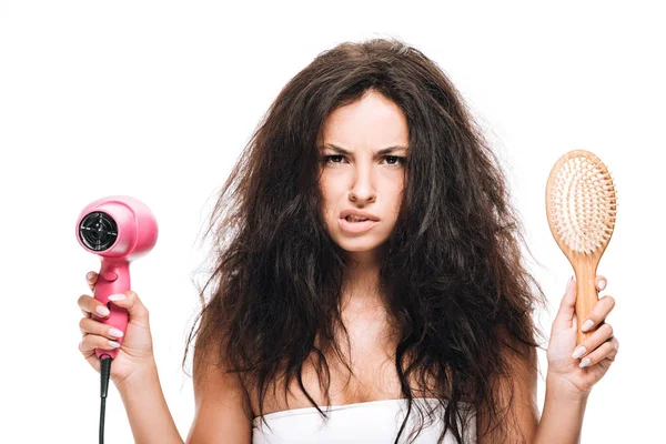 Confused brunette beautiful woman styling curly hair with pink hairdryer and comb isolated on white — Stock Photo