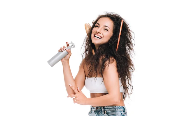 Mulher morena feliz com pentes no cabelo ondulado unruly segurando spray isolado no branco — Fotografia de Stock