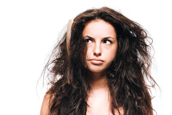 Confused brunette woman with hairbrush in unruly curly hair isolated on white — Stock Photo