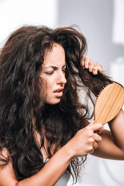 Confused brunette woman styling unruly curly hair with hairbrush — Stock Photo