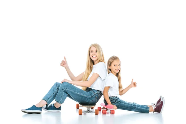 Happy mother and daughter sitting on penny boards and showing thumbs up on white — Stock Photo