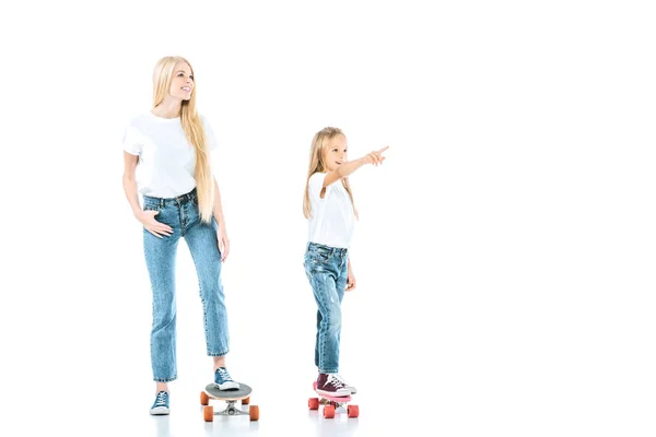 Mãe feliz montando penny board perto filha apontando com o dedo isolado no branco — Fotografia de Stock