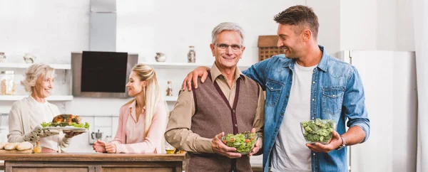 Panoramic shot of smiling grandfather and father hugging and holding bowls with salad and broccoli in Thanksgiving day — Stock Photo