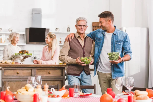 Nonno e padre sorridenti che abbracciano e tengono ciotole con insalata e broccoli nel giorno del Ringraziamento — Foto stock