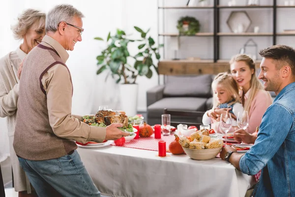 Family members sitting at table and grandfather holding plate with turkey in Thanksgiving day — Stock Photo