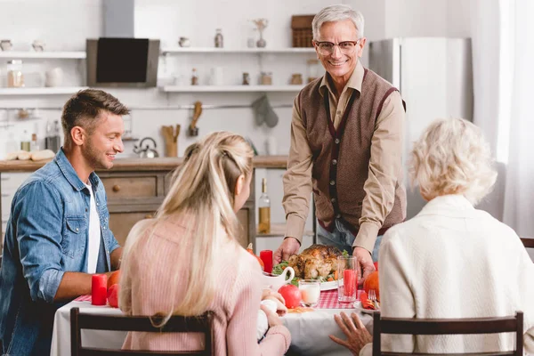 Membres de la famille assis à la table et grand-père tenant assiette avec dinde le jour de Thanksgiving — Photo de stock