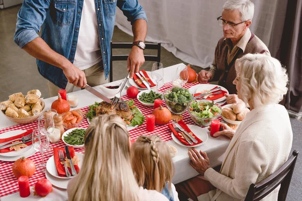 Vista de ángulo alto de los miembros de la familia sentados en la mesa y el padre de corte sabroso pavo en el día de Acción de Gracias - foto de stock