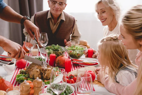 Vista recortada de los miembros de la familia sentados a la mesa y el padre de corte sabroso pavo en el día de Acción de Gracias - foto de stock