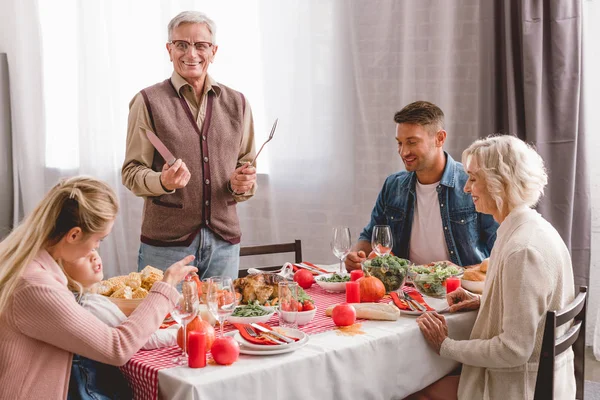 Miembros de la familia sentados a la mesa y el abuelo sosteniendo cubiertos en el día de Acción de Gracias - foto de stock