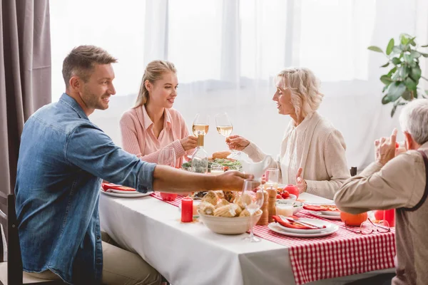 Miembros de la familia sonrientes sentados a la mesa y tintineando con copas de vino en el día de Acción de Gracias - foto de stock