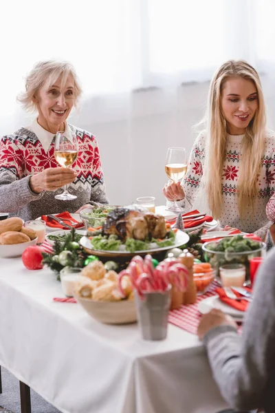 Miembros de la familia sentados a la mesa y sosteniendo copas de vino en Navidad - foto de stock