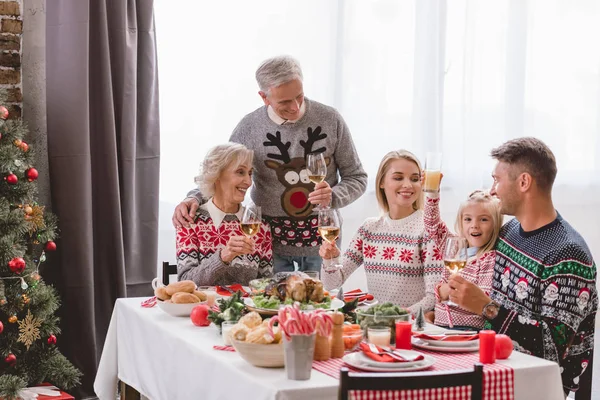 Miembros de la familia sentados a la mesa y sosteniendo copas de vino en Navidad - foto de stock