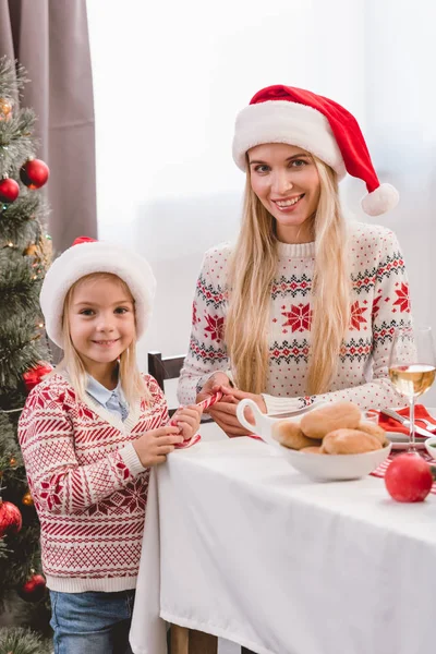 Madre e hija en suéteres y sombreros de santa sonriendo y mirando a la cámara en Navidad - foto de stock