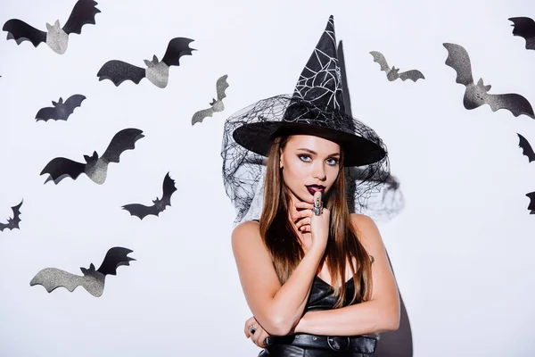 Mädchen in schwarzem Hexenhalloween-Kostüm blickt in die Kamera neben weißer Wand mit dekorativen Fledermäusen — Stockfoto