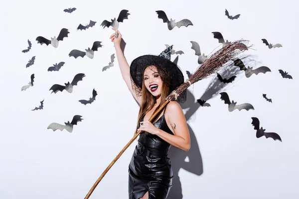 Девушка в черной ведьме Хэллоуин костюм с метлой рядом с белой стеной с декоративными летучих мышей — стоковое фото