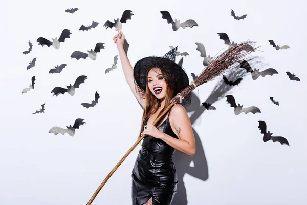 Girl in black witch Halloween costume with broom near white wall with decorative bats — Stock Photo