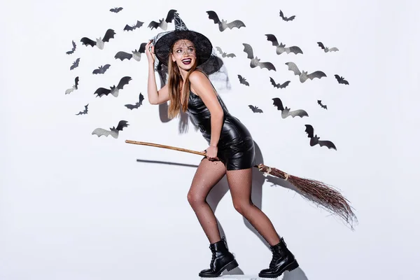 Glückliches Mädchen in schwarzem Hexenhalloween-Kostüm auf Besen neben weißer Wand mit dekorativen Fledermäusen — Stockfoto